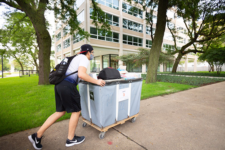 A student transports his belongings to Mies Campus housing wearing summer clothing and his all-important protective mask.